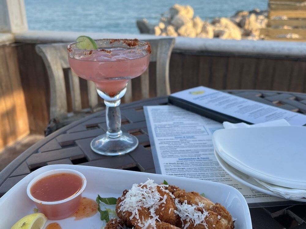 A plate of coconut shrimp with sauce and a lemon wedge sits on a patio table next to a pink margarita, menu, and stacked plates, overlooking a rocky shore and calm ocean under a blue sky.