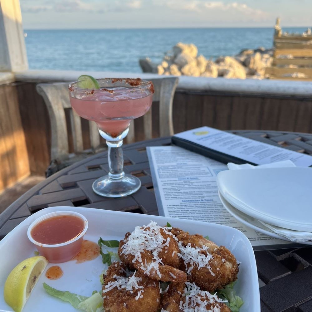 A plate of coconut shrimp with sauce and a lemon wedge sits on a patio table next to a pink margarita, menu, and stacked plates, overlooking a rocky shore and calm ocean under a blue sky.