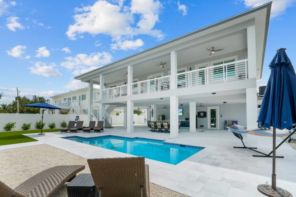 Modern two-story white house with a large balcony, outdoor ceiling fans, pool, patio furniture, lounge chairs, a hammock, and blue umbrellas under a partly cloudy sky. The setting is clean and inviting.