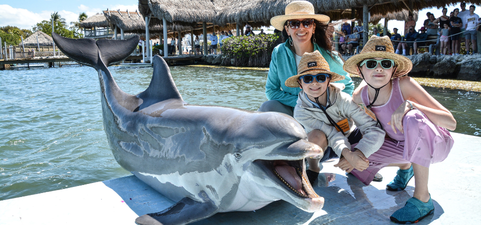 A woman and two children in sun hats pose with a smiling dolphin on a dock at a tropical marine park, with thatched-roof buildings and a crowd of people in the background.