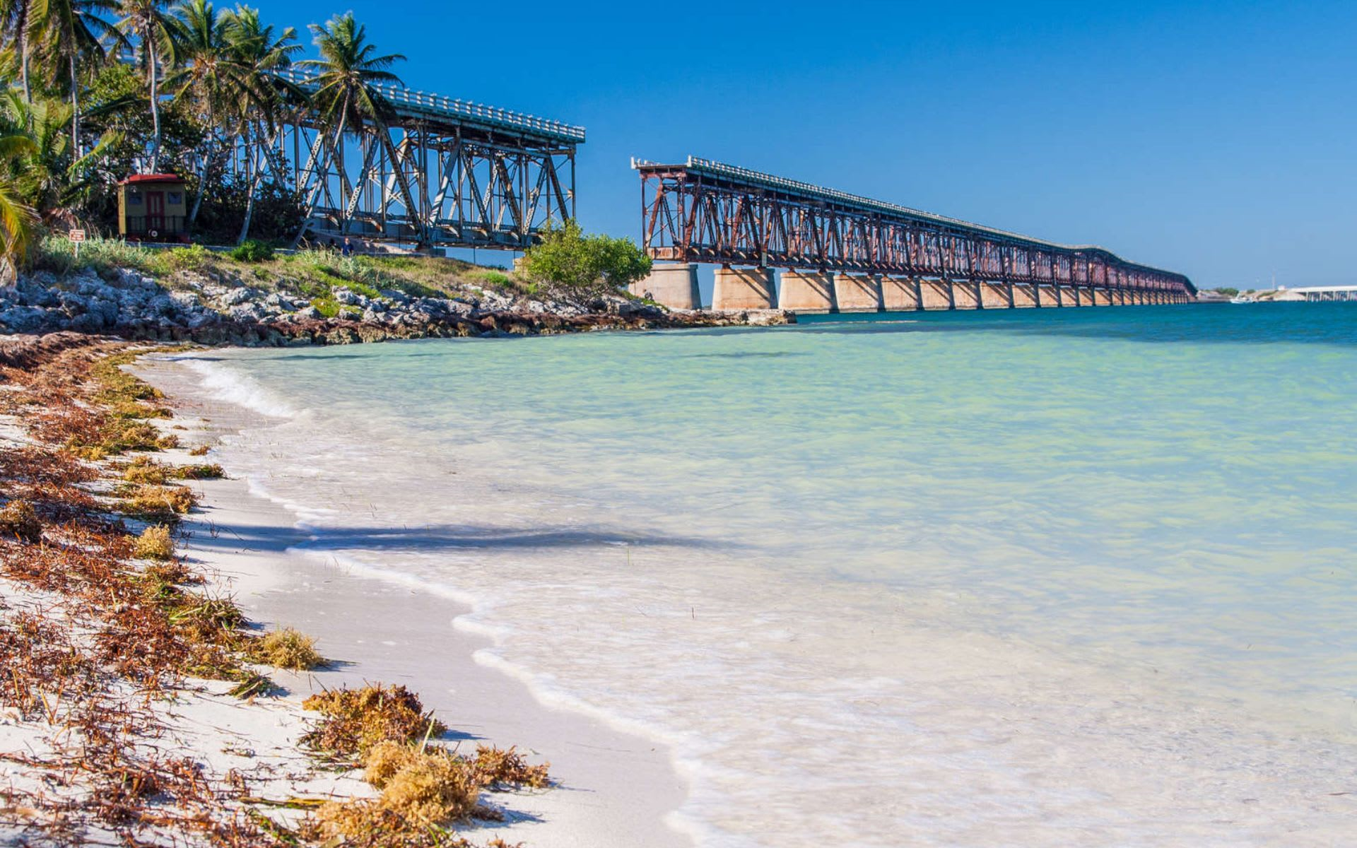 Sombrero beach bridge: a long, rusty bridge stretches over clear turquoise water near a sandy beach lined with seaweed and palm trees, under a bright blue sky.