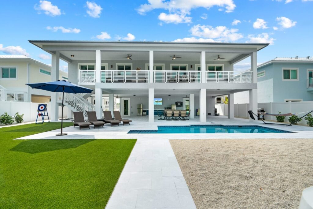 Modern two-story house with large balcony, pool, lounge chairs, and umbrella in backyard. The yard has green grass, sand area, spacious patio, and is surrounded by other similar houses under a blue sky with clouds. (luna light: marathon fl vacation rental | villa paraiso)