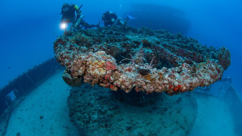 Two scuba divers explore a large, coral-encrusted shipwreck on the ocean floor, shining their flashlights on the structure as fish swim nearby in the blue water.