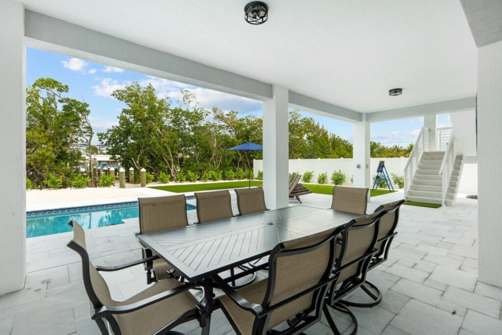 A covered patio with a metal dining table and eight chairs overlooks a backyard with a swimming pool, lounge chairs, green lawn, and trees in the background. Stairs lead to an upper level.