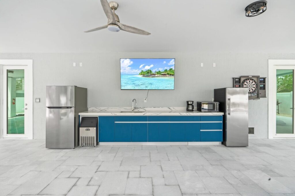 Modern outdoor kitchen with blue cabinets, stainless steel appliances, a wall-mounted tv displaying a beach scene, a ceiling fan, dartboard, and light gray tiled floor.