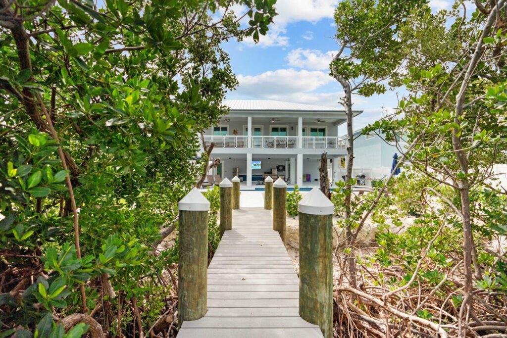 A wooden dock surrounded by greenery leads to a two-story white house with a large balcony and palm trees, under a blue sky with scattered clouds.