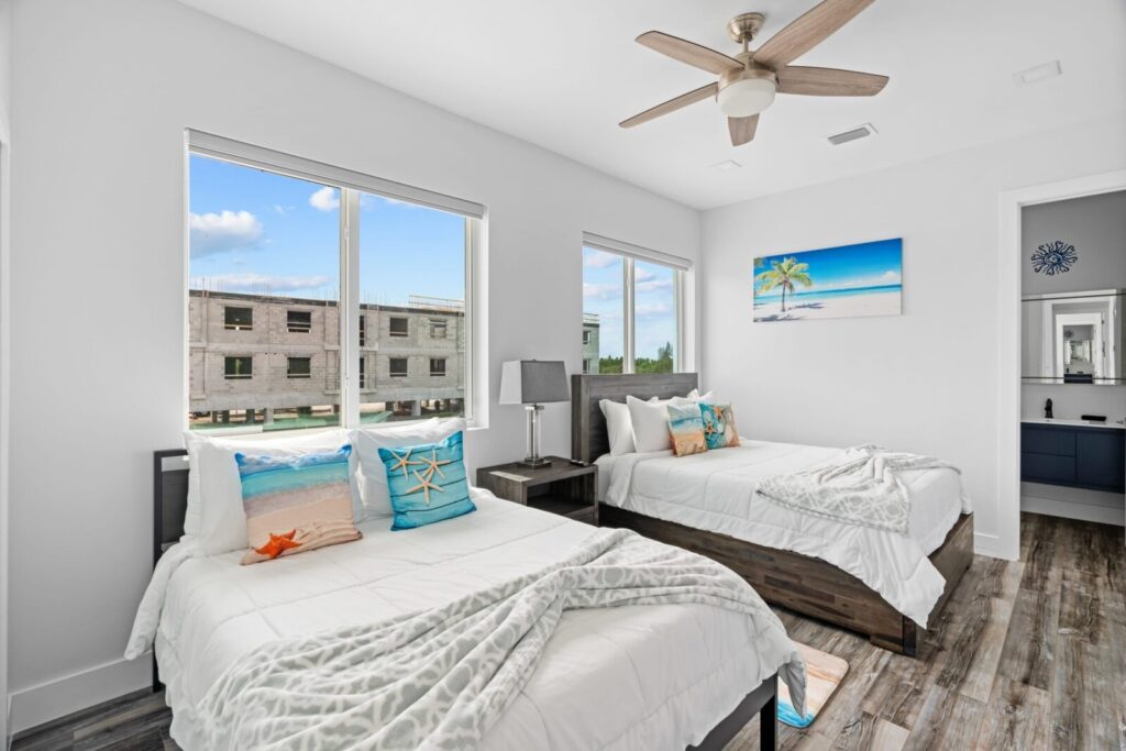 Bright bedroom with two double beds, white bedding, coastal-themed pillows, a ceiling fan, large windows, and a beach painting on the wall. An ensuite bathroom is visible in the background.