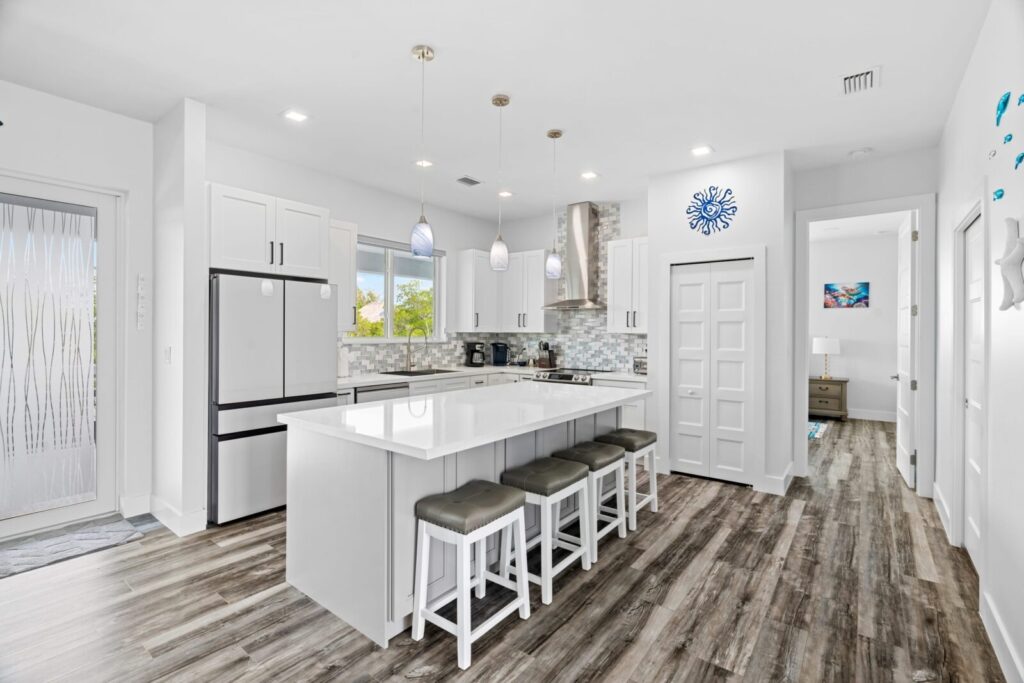 Modern white kitchen with an island, four stools, stainless steel appliances, pendant lighting, and a mosaic backsplash. Wood-style flooring and a bedroom are visible through an open doorway.