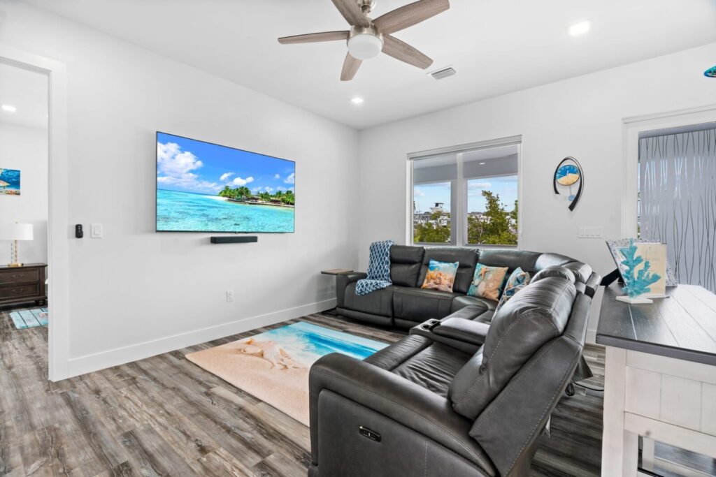 Modern living room with a gray sectional sofa, beach-themed decor, wood floor, a wall-mounted tv displaying a tropical ocean scene, and large windows letting in natural light.
