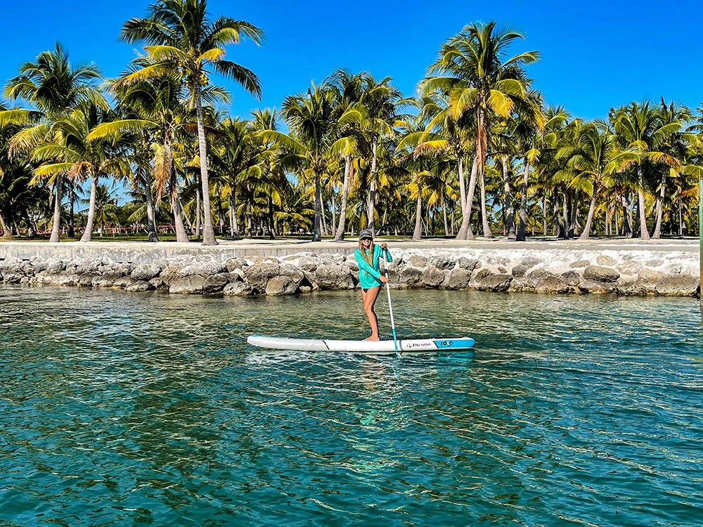 A person in a teal shirt and shorts enjoys paddleboarding in marathon fl on clear blue water near a rocky shore lined with tall palm trees under a bright blue sky.