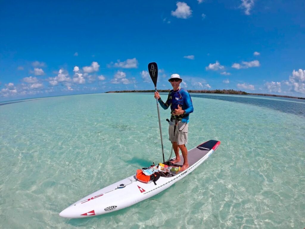 A person wearing a hat, sunglasses, and blue shirt stands on a paddleboard in clear, shallow turquoise water under a bright blue sky in marathon fl, giving a thumbs-up—perfect day for paddleboarding in marathon fl.