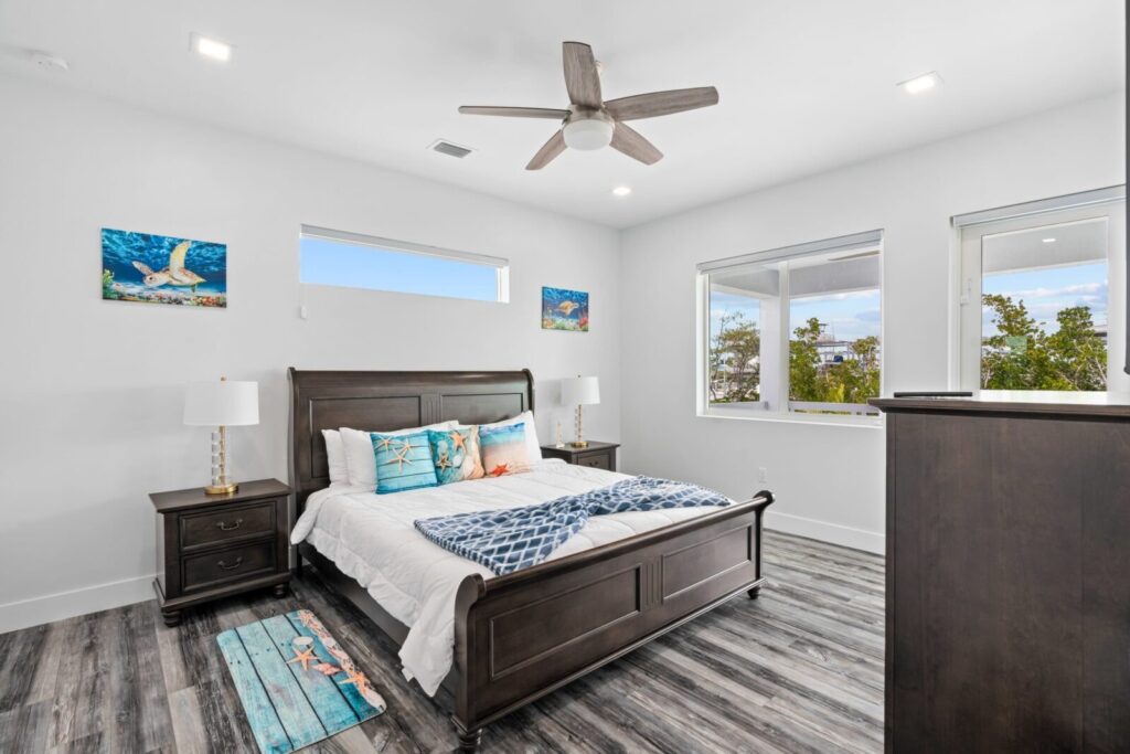 A bright bedroom with a dark wood bed, white bedding, blue throw blanket, and beach-themed pillows. There are two nightstands with lamps, ocean-themed wall art, large windows, and a ceiling fan. The floor is wood-patterned.