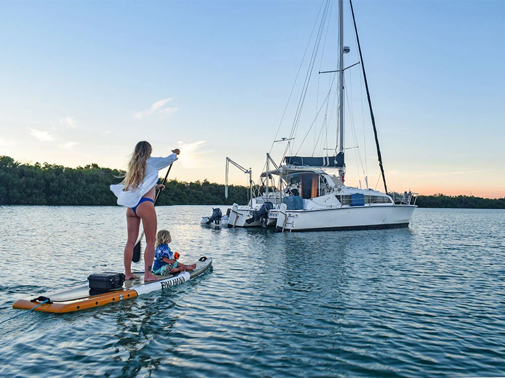 A woman paddleboarding in marathon fl with a young child seated in front of her glides across calm water toward an anchored sailboat, surrounded by trees and a clear evening sky.