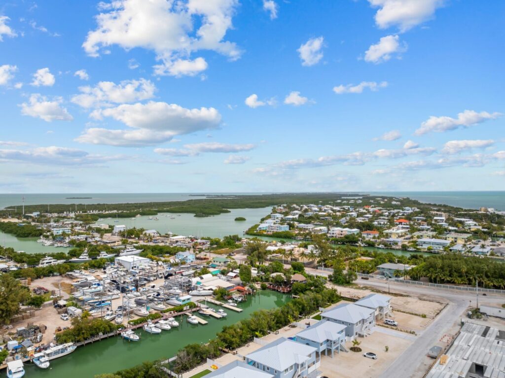 Aerial view of a coastal town with marinas, boats docked along a canal, houses, lush greenery, and blue skies with scattered clouds above.