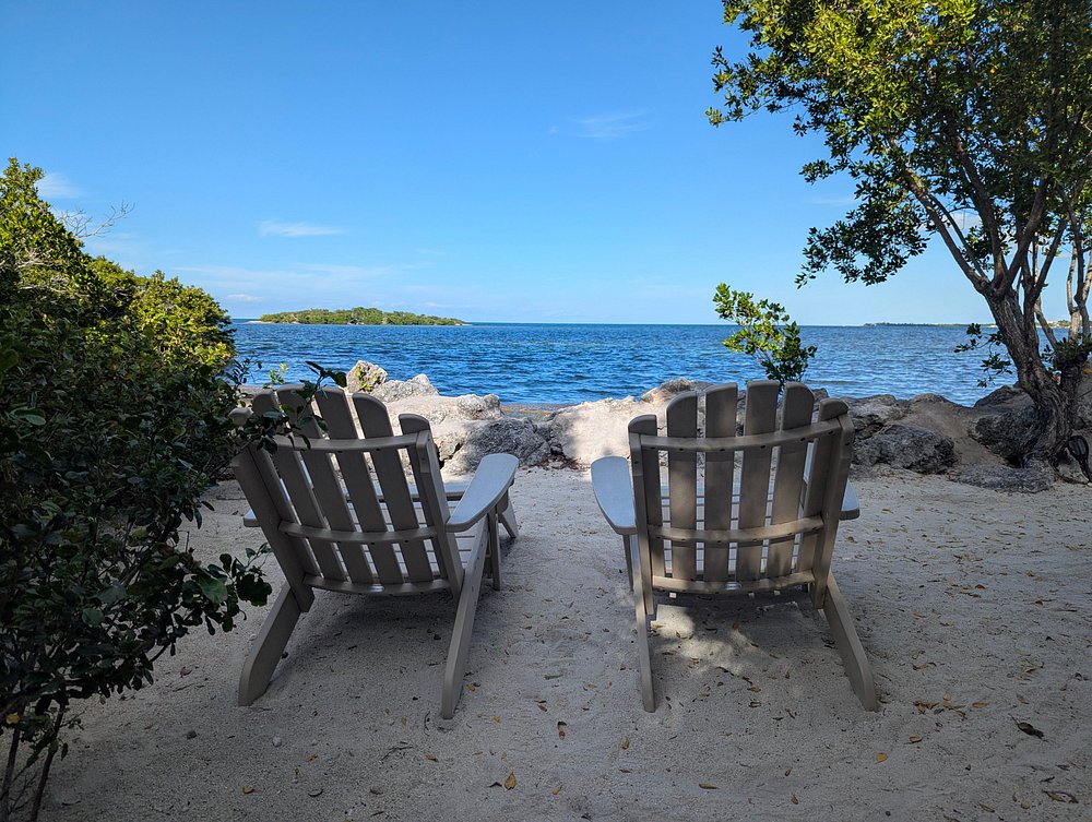 Two wooden lounge chairs sit on a sandy beach facing a clear blue sea, surrounded by green trees and bushes, with a small island visible in the distance under a bright blue sky.