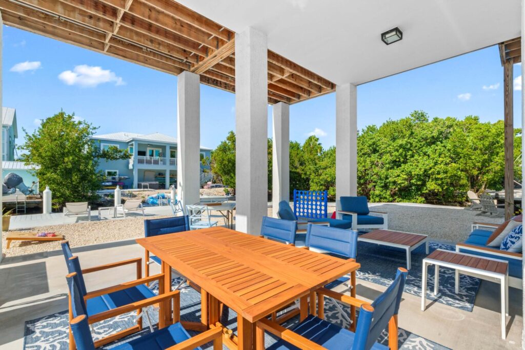 A covered outdoor patio with a wooden dining table and blue chairs, additional lounge seating, a large connect four game, and surrounding greenery on a sunny day. Modern houses are visible in the background.