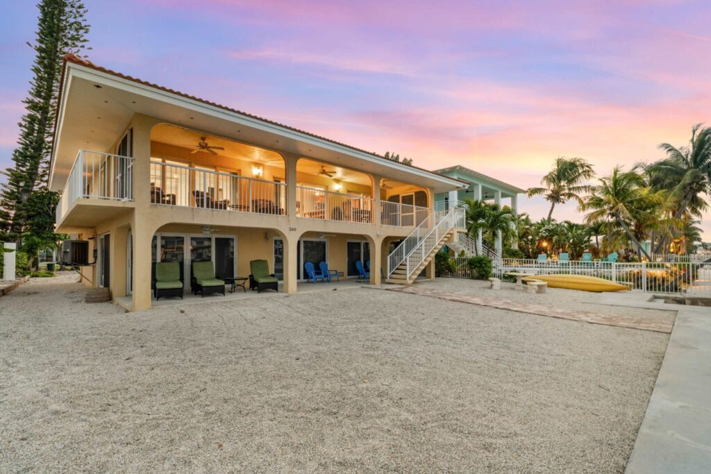 Two-story beach house with large balcony, outdoor seating, and stairway. Surrounded by palm trees and a sandy yard at sunset, with glowing indoor lights and a vibrant sky in the background.