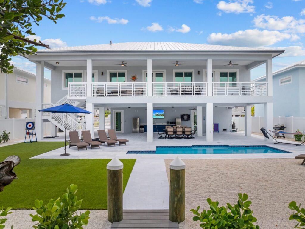 Modern two-story white house with a large balcony, outdoor lounge chairs, swimming pool, green artificial lawn, patio dining area, and blue sky with scattered clouds. White fence and palm trees surround the backyard.