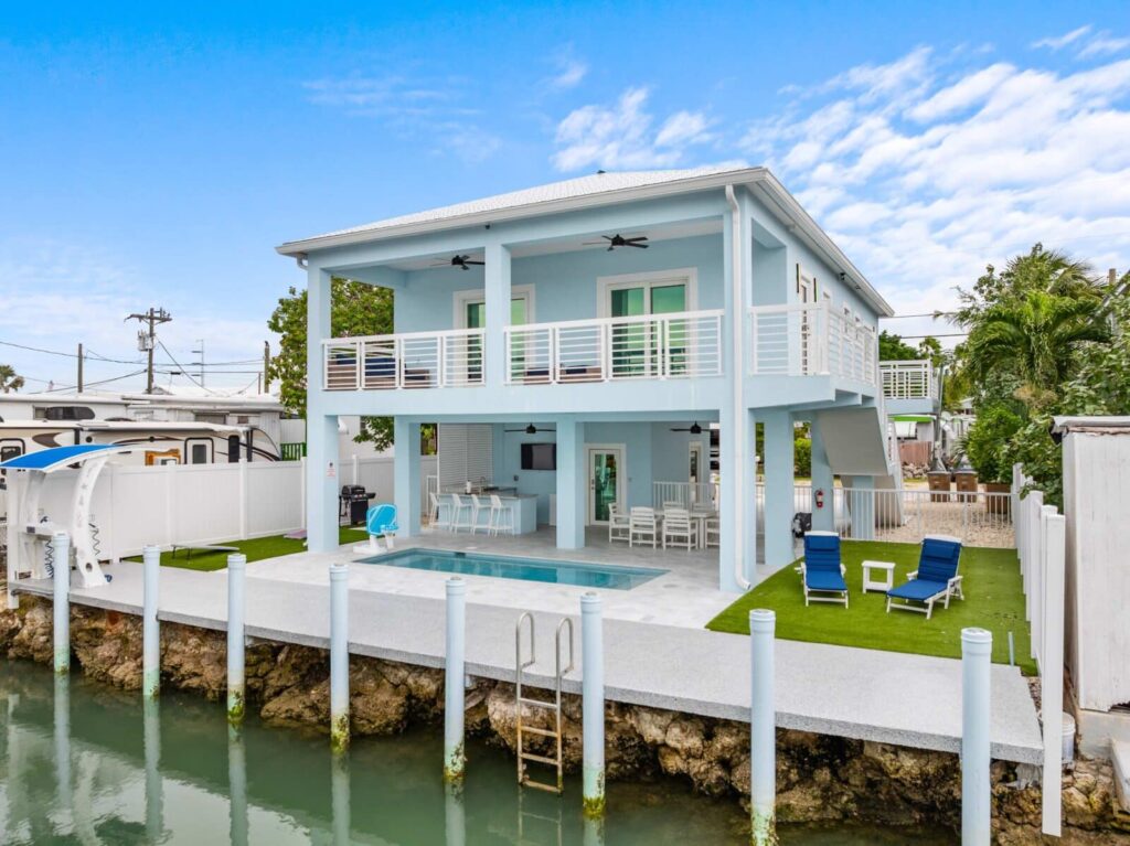 Vacation rental insurance 101 for marathon, fl owners (2026 guide) 4 A modern two-story light blue house with a large balcony, pool, lounge chairs, and canal access with a ladder, set in a tropical area under a partly cloudy sky.