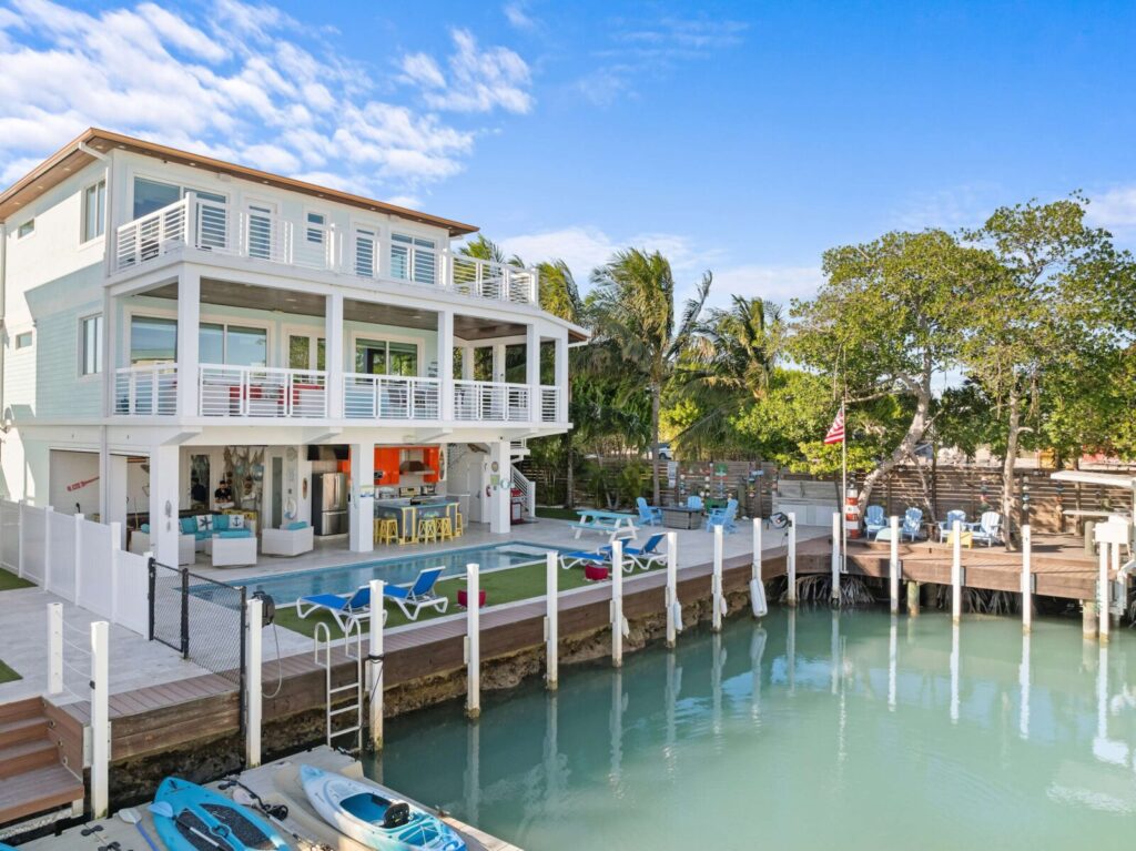 A modern three-story waterfront house with large balconies, a pool, outdoor seating, kayaks by the dock, and clear blue skies. Palm trees and a wooden deck with chairs are in the background.