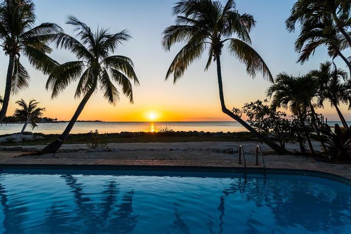 Sunset over a calm ocean, viewed from behind a swimming pool. Palm trees frame the scene, with the sky glowing orange and blue above the tranquil water.