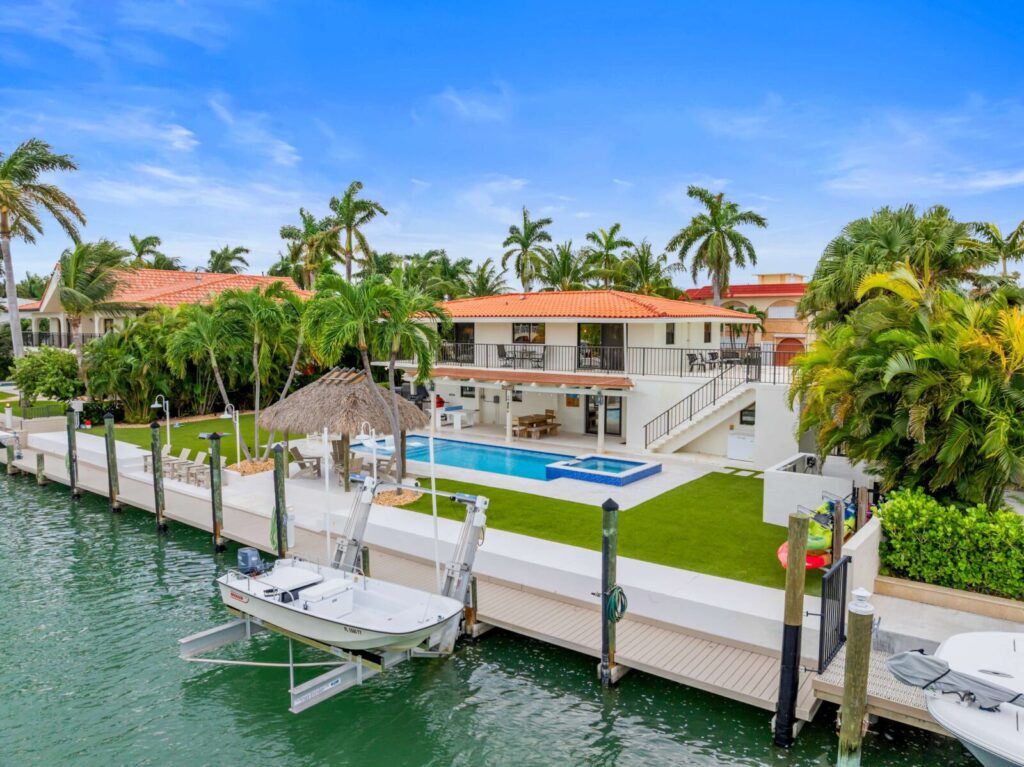 Waterfront villa with a red-tiled roof, pool, hot tub, and thatched gazebo, surrounded by palm trees. A boat is docked by the green lawn along the canal under a blue sky.