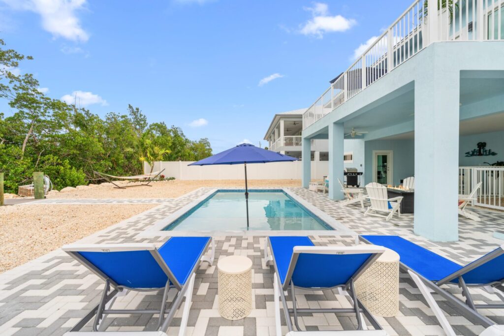 A backyard with a rectangular pool, blue lounge chairs, and a large blue umbrella. Theres patio seating under a covered area, a hammock in the background, and trees and a fence surrounding the space.