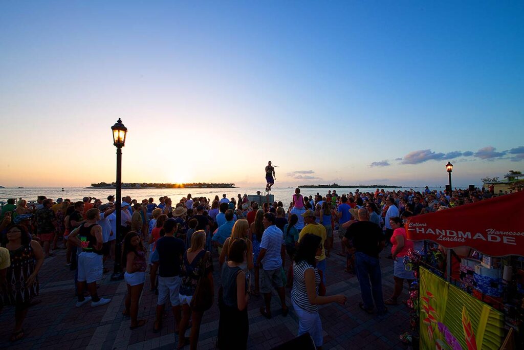 Marathon vs key west: which florida keys destination is right for you? 5 A large crowd gathers on a boardwalk at sunset, watching a street performer elevated above the group. The sky is clear, and there are street lamps, with the ocean and clouds in the background. A red vendor tent is at the right.