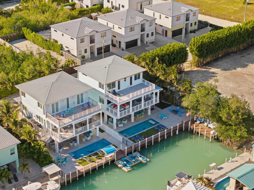 Vacation rental insurance 101 for marathon, fl owners (2026 guide) 2 Aerial view of waterfront houses with docks, swimming pools, patios, and boats along a turquoise canal. Several modern, two-story homes are surrounded by palm trees and greenery.