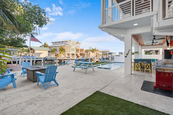 Vacation rental insurance 101 for marathon, fl owners (2026 guide) 3 Spacious outdoor patio with blue adirondack chairs around a firepit, picnic tables, a pool, outdoor kitchen, and canal views. Palm trees and houses line the waterfront, with clear blue skies above.