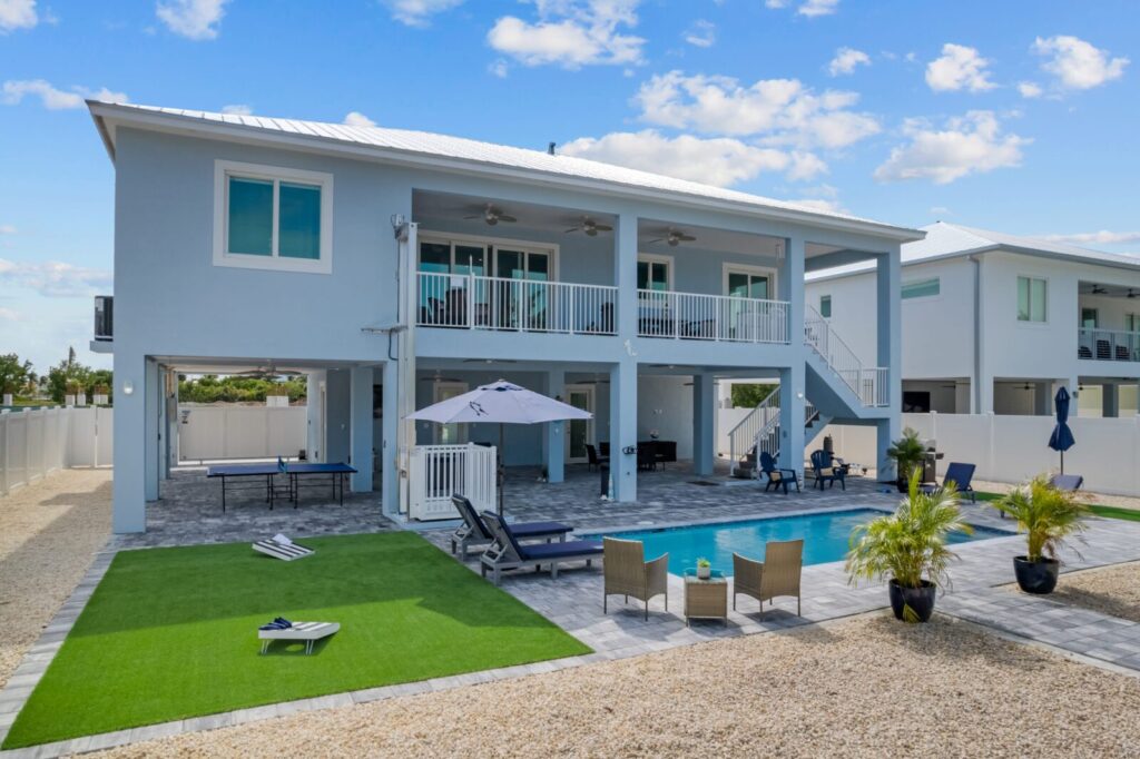 A modern two-story house with light blue siding, a large balcony, and a covered patio beside a rectangular swimming pool, outdoor lounge chairs, and small palm trees in a spacious backyard.