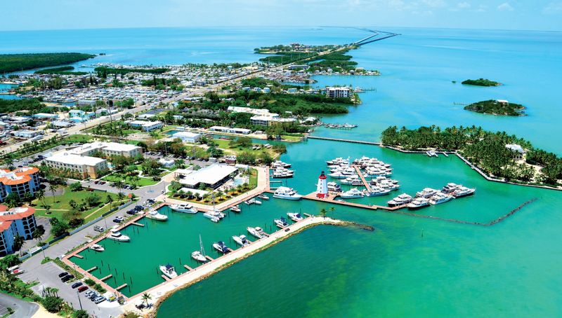 Aerial view of a marina with boats docked in turquoise water, surrounded by buildings, palm trees, and roads leading into the distance over the ocean with small islands visible.