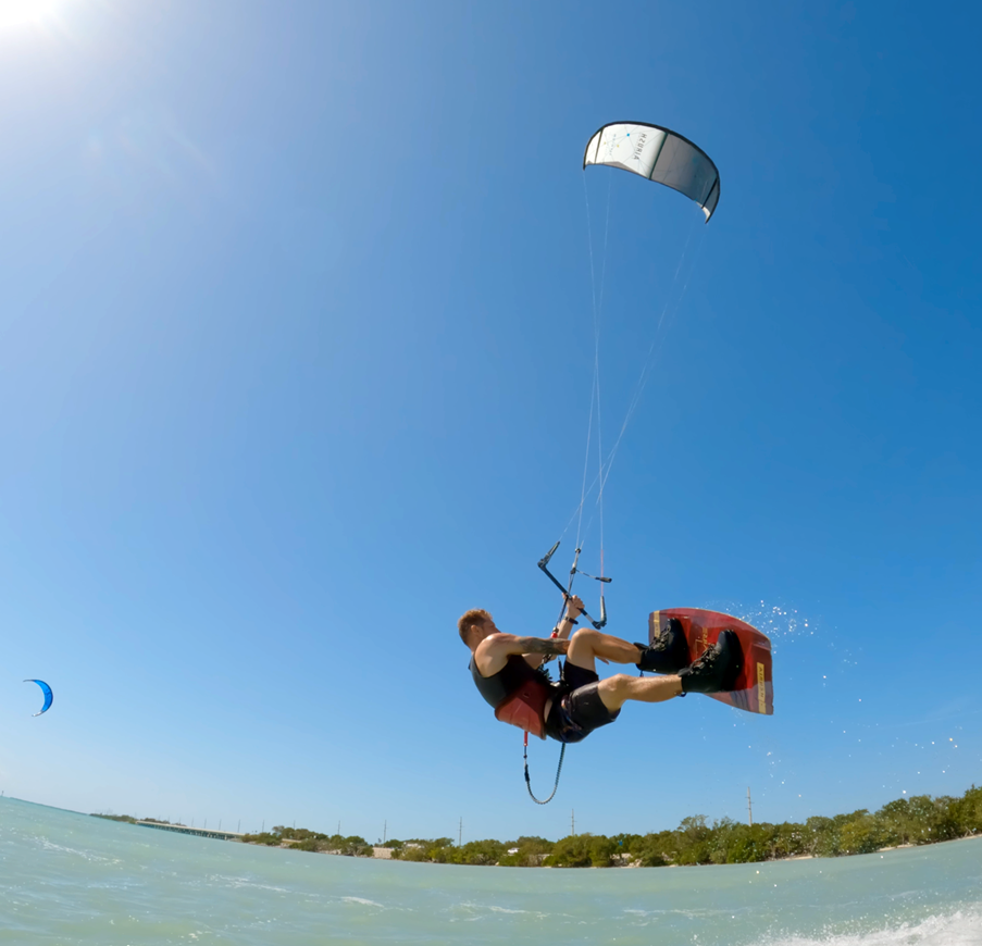 A person is kiteboarding, catching air above the water with a blue sky overhead. They are holding onto the control bar while the kite pulls them up, and the shoreline with trees is visible in the background.