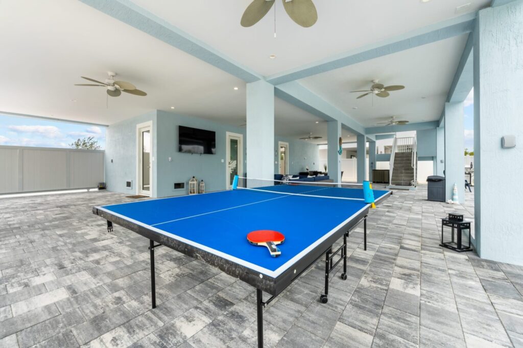 A blue ping pong table with paddles and balls is set up on a covered patio with ceiling fans, gray tiled flooring, and light blue walls. Outdoor furniture and a tv are visible in the background.