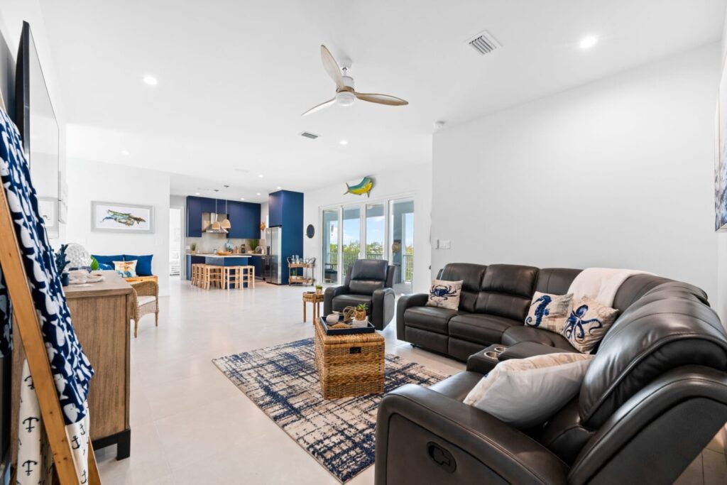 Modern, open-concept living room with black leather sofas, a patterned rug, and a ceiling fan. The space extends into a kitchen with blue cabinetry and large windows leading to an outdoor view. Decor includes nautical accents.