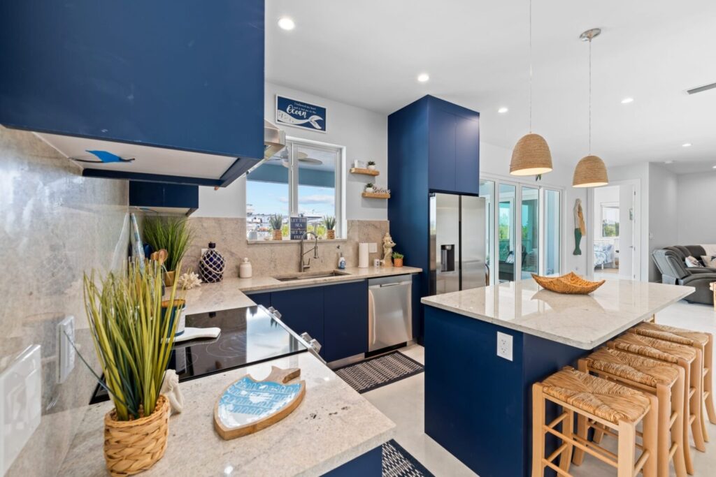 Modern kitchen with navy blue cabinets, white granite countertops, an island with wicker stools, pendant lights, stainless steel appliances, and decorative plants, with a view of a bright living area and large windows.