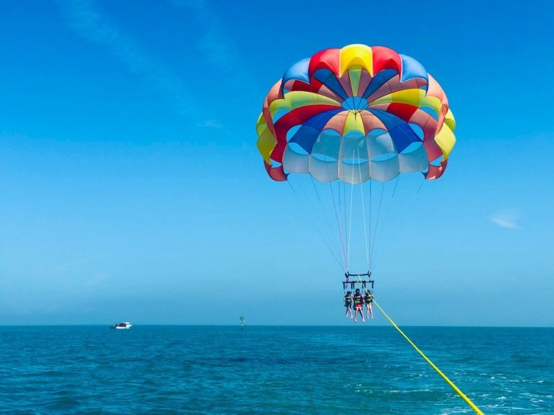Two people parasailing high above the blue ocean under a colorful parachute, with a clear blue sky and a distant boat on the water.
