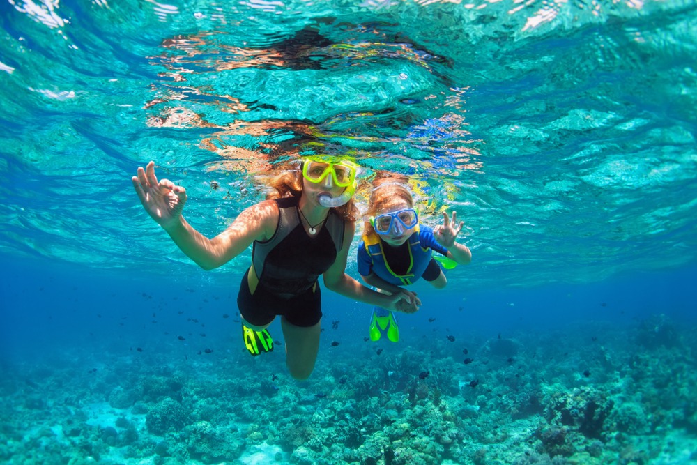 Two people wearing snorkel gear and swim fins float underwater above a coral reef, making ok hand signals in clear blue water with sunlight streaming through the surface.