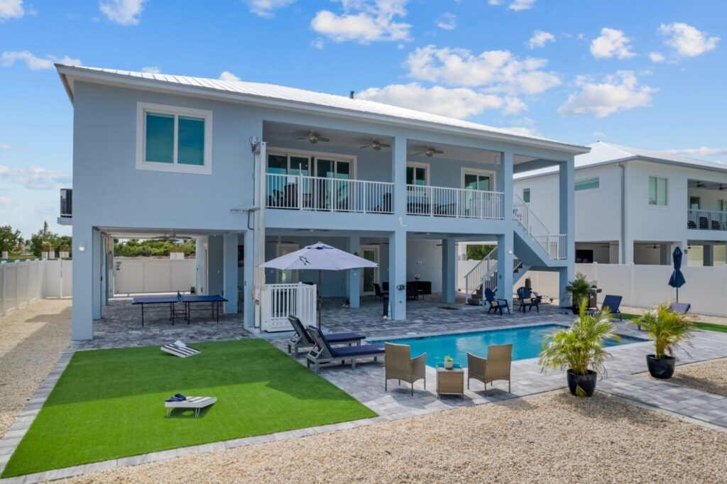 A modern two-story house with a large balcony overlooks a backyard featuring a swimming pool, lounge chairs, a patio set, green artificial turf, and palm plants under a blue sky with scattered clouds.