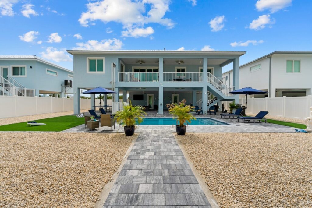 A modern two-story house with balconies overlooks a backyard featuring a swimming pool, lounge chairs, patio seating, and potted plants, all set on a stone walkway and gravel yard under a blue sky with scattered clouds.