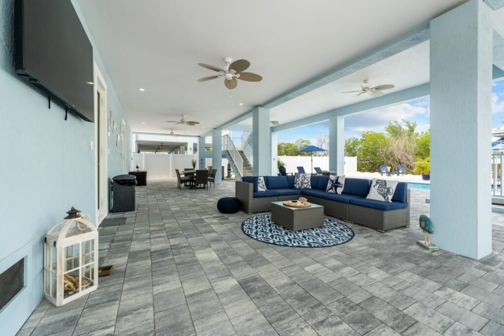 Spacious covered patio with blue walls, gray tile flooring, ceiling fans, outdoor seating area with blue cushions, tv, lantern, and a view of a pool and greenery in the background.