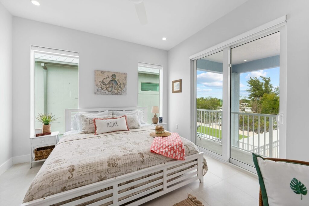 Bright, modern bedroom with a large bed, nautical-themed bedding, and two windows. Glass doors open to a balcony with a view of trees and sky. Decor includes a relax pillow, plant, and wall art above the bed.