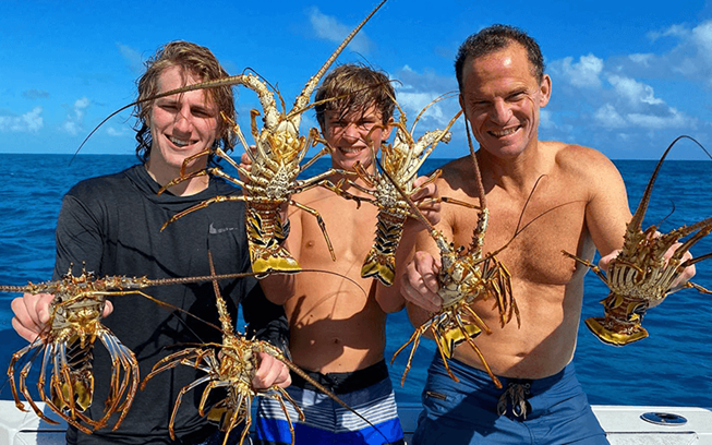Three smiling people on a boat hold up several large lobsters with the ocean and blue sky in the background. They appear happy and are wearing swimsuits.