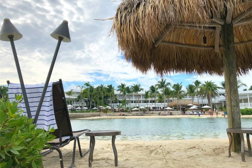 A sandy beach with lounge chairs, a striped towel, and a straw umbrella overlooks a calm lagoon—an inviting spot where mistakes first-time florida keys visitors make often include forgetting sun protection under the partly cloudy sky.