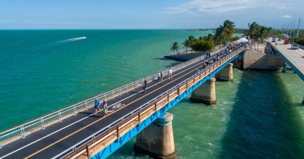 Marathon vs key west: which florida keys destination is right for you? 3 Aerial view of a long pedestrian bridge over turquoise ocean water; people are walking and cycling, with palm trees and a road with cars to the right under a clear blue sky.