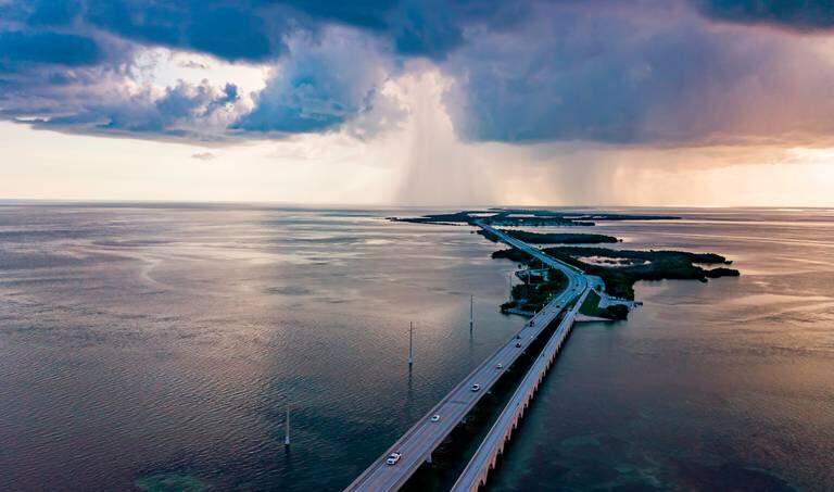 A long bridge stretches over calm blue water toward distant islands in the florida keys. Cars drive across as dark storm clouds and rain gather on the horizon, while sunlight breaks through on the right, evoking a dramatic summer scene.
