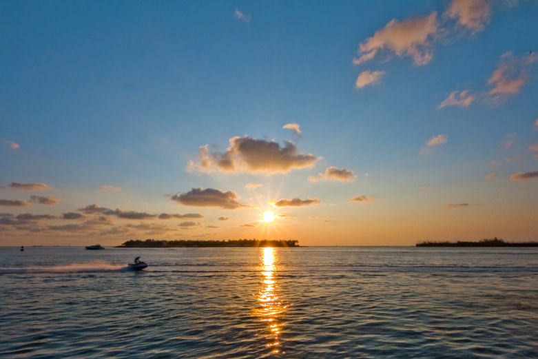 A jet ski moves across calm water at sunset in the florida keys, with the sun low on the horizon, casting reflections on the water and illuminating scattered clouds—a perfect summer scene with small islands and boats in the distance.