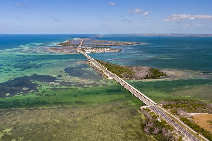 A long bridge stretches across clear blue and turquoise waters, connecting small islands covered in greenery. The scene is viewed from above under a sunny sky with scattered clouds.