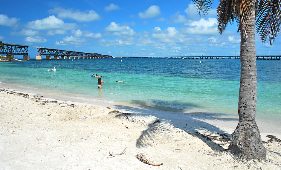 Marathon vs key west: which florida keys destination is right for you? 6 A sandy beach with turquoise water, people swimming, and an old bridge stretching across the sea in the background. A palm tree casts a shadow on the sand under a sunny sky with scattered clouds.