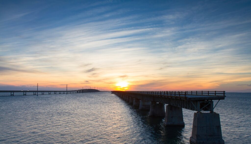 A long bridge extends across a large body of water toward the horizon, with the sun setting and colorful clouds filling the sky. The scene is peaceful, with warm orange and blue hues reflecting on the water.